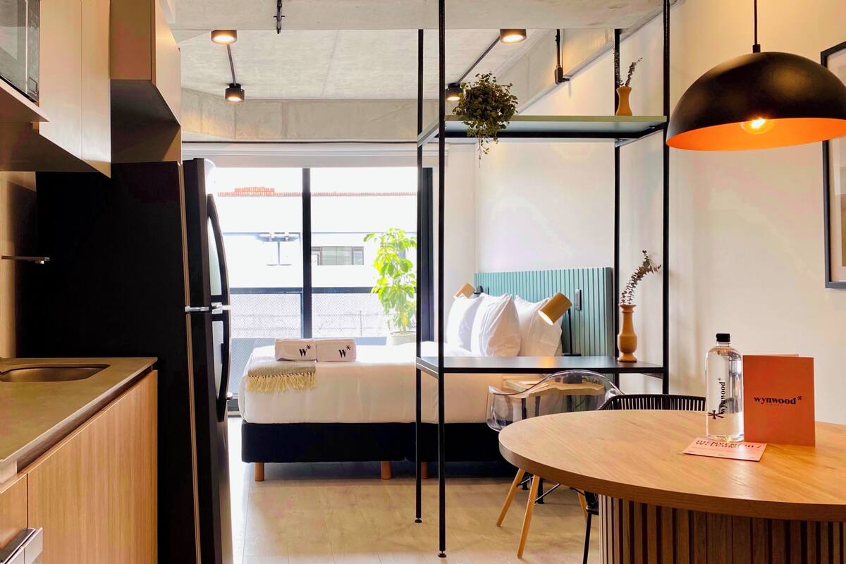 Une chambre d’hôtel moderne avec un lit, une table ronde en bois, un coin cuisine et une grande fenêtre.