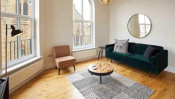 Een moderne woonkamer met een groene bank, een ronde salontafel en een roze fauteuil. Er zijn twee grote ramen met uitzicht op gebouwen.