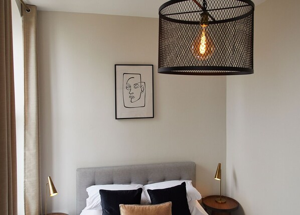 Une chambre à coucher avec une tête de lit recouverte d’un tissu gris, un lit avec une literie blanche et noire, une œuvre d’art encadrée au mur et un luminaire suspendu avec une ampoule visible.