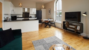 Een moderne keuken met witte kasten, een zwarte koelkast, en een eethoek met een ronde tafel en stoelen. Er is een groot raam dat natuurlijk licht binnenlaat, en een flatscreen televisie aan de muur bevestigd.