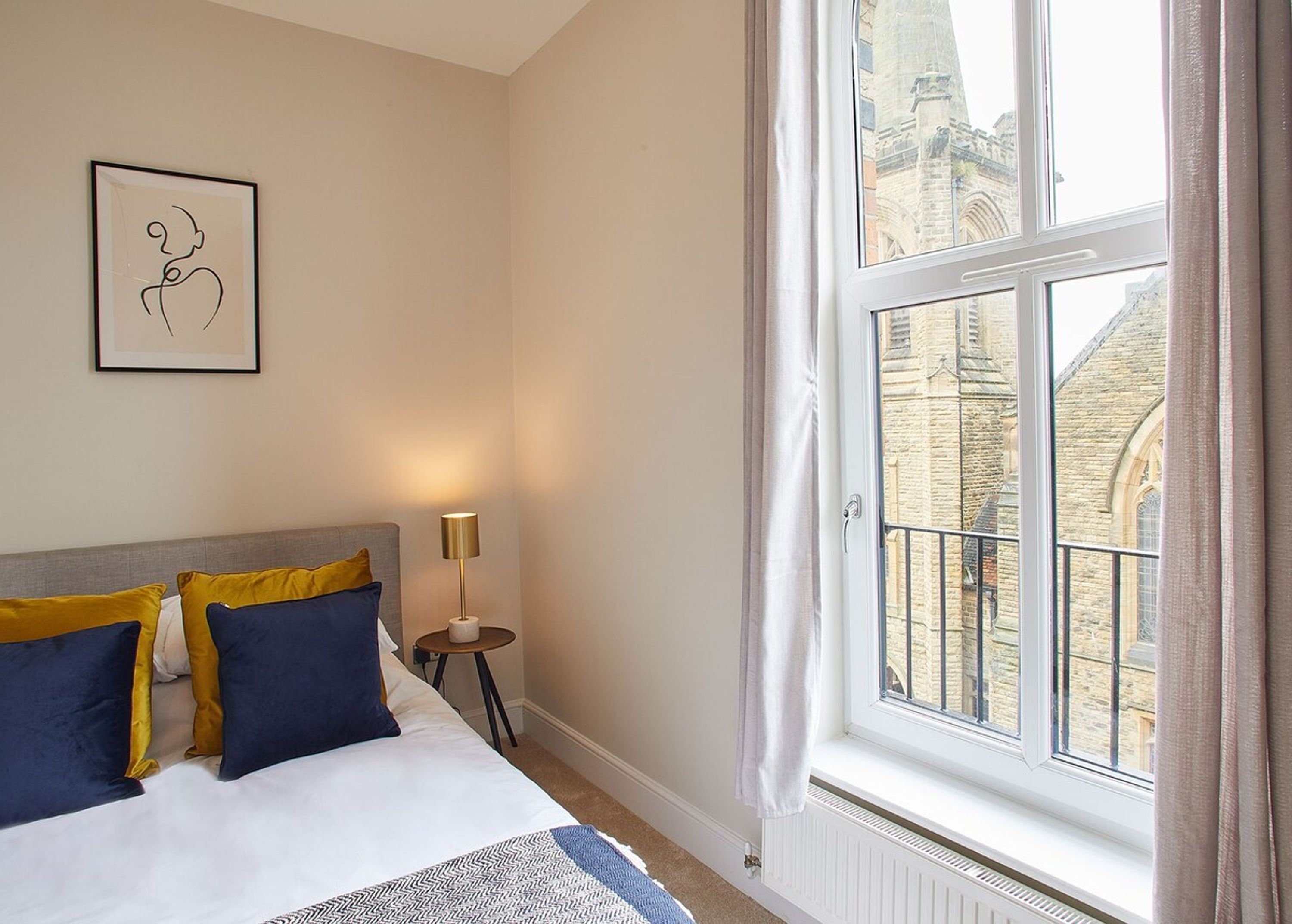 A hotel room with a bed, a bedside table with a lamp, a window with curtains, and a framed picture on the wall.