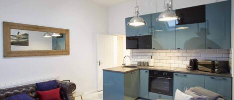A modern kitchen with blue cabinets, a built-in oven, and a microwave. There is a wooden-framed mirror on the wall, a brown leather sofa with red and blue cushions, and a grey armchair.