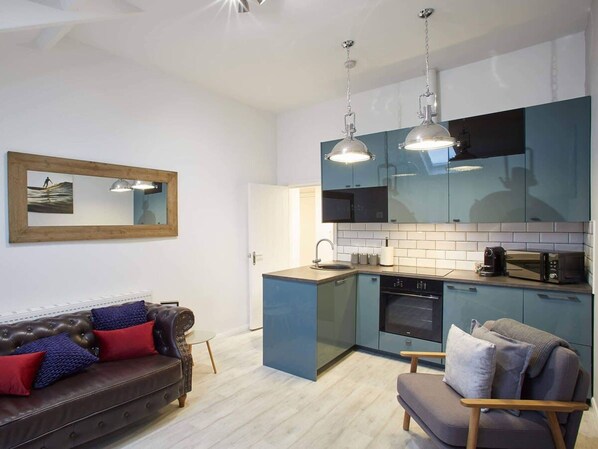 A modern kitchen with blue cabinets, a built-in oven, and a microwave. There is a brown leather sofa with red and blue cushions, and a wooden chair with a grey cushion. A wooden-framed mirror hangs on the wall.