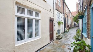 A narrow stone-paved alleyway between buildings with white and blue facades, a brown door, and potted plants. - Host Stay Threadneedle Cottage (Whitby)