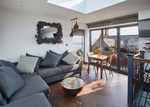 A modern living room with a grey sofa, patterned cushions, a coffee table, and a large mirror. There is a balcony with a view of buildings and a dining area with a table and chairs. - Host Stay The Nook (Saltburn-by-the-Sea)