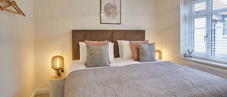Une chambre à coucher avec un grand lit, une tête de lit en bois, un luminaire suspendu et une fenêtre avec des stores.