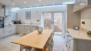 Cocina moderna con comedor, que incluye una mesa de madera, armarios blancos y vistas a un muro de ladrillo exterior a través de un ventanal.