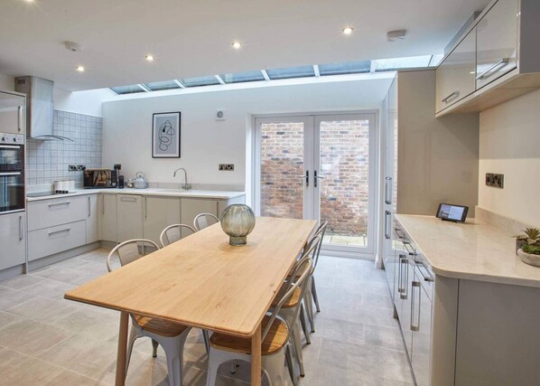 A modern kitchen with a dining area, featuring a wooden table, white cabinets, and a view through a glass door to an outdoor brick wall. - Host Stay Hoggarths Retreat (Whitby)