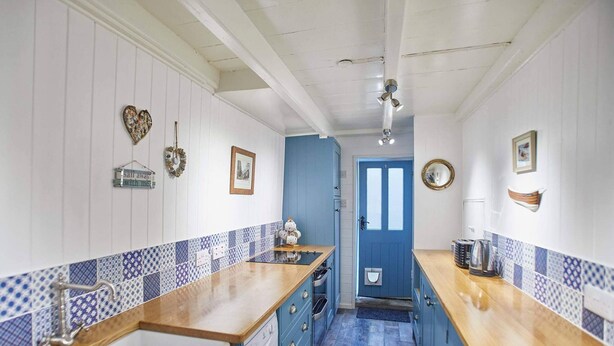 A narrow kitchen with blue and white tiled backsplash, wooden countertops, and a blue door.
