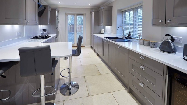 Cocina moderna con armarios grises, encimera blanca y estufa de acero inoxidable. Hay una zona de bar con una silla alta y taburetes.