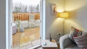 Una sala de estar acogedora con una puerta corrediza de cristal que da a un patio de piedra, una lámpara y un cojín en un sofá.