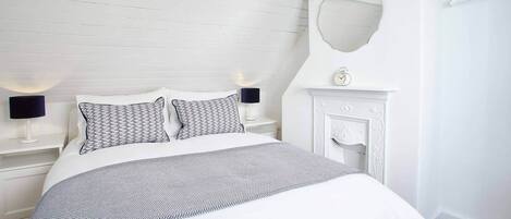 A neatly arranged bedroom with a bed, two bedside tables with lamps, a mirror, and a fireplace.