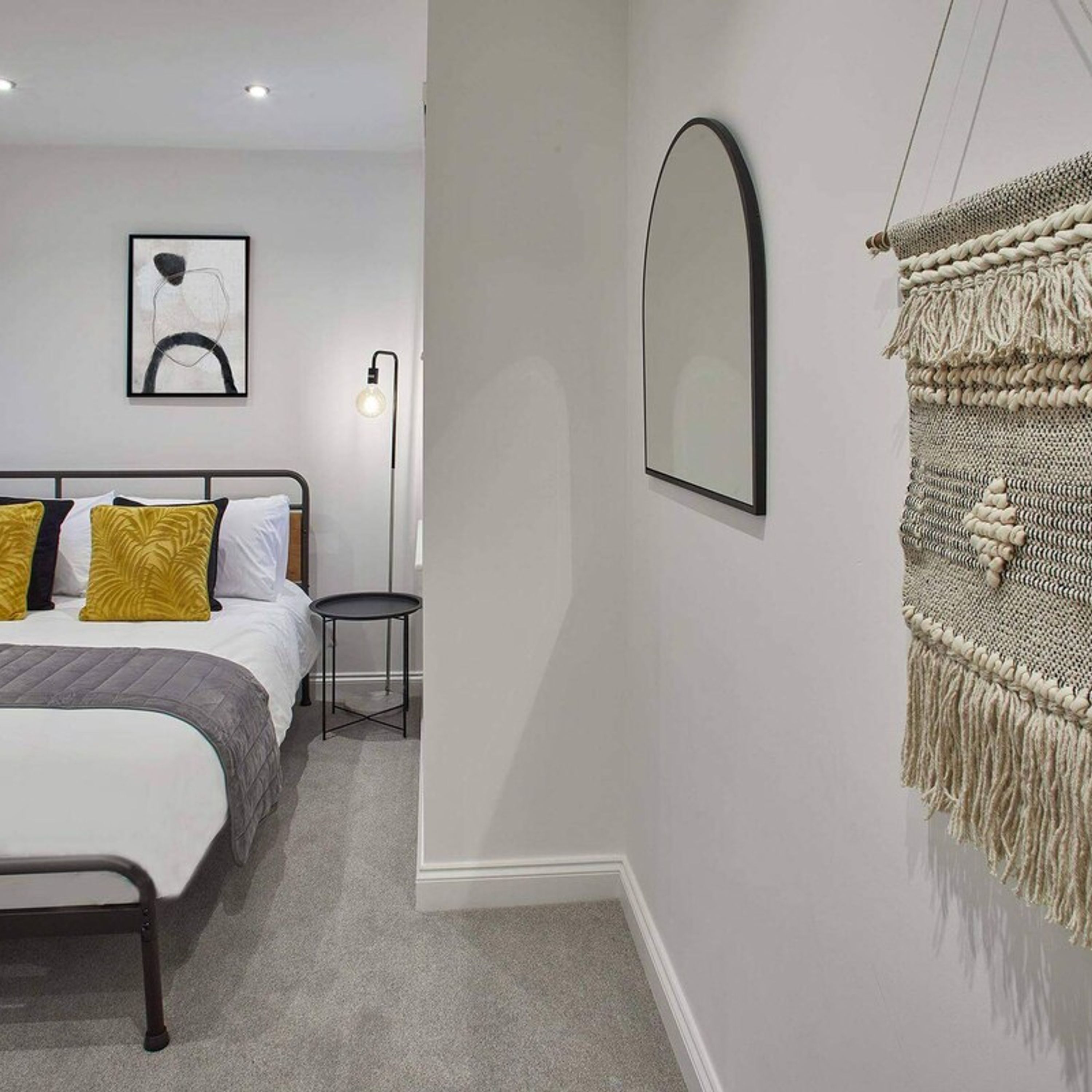 A bedroom with a bed, bedside table, lamp, artwork, and a macrame wall hanging.