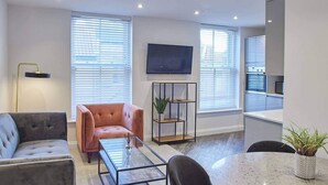 A modern living room with a grey sofa, a pink armchair, a glass coffee table, and a round dining table. There is a TV mounted on the wall, a floor lamp, and a plant on a shelf. - Host Stay Apartment Two Hudsons Yard House (Whitby)