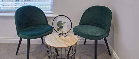 A corner room with two green chairs, a small round table, and a window with blinds.