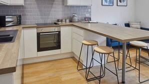 Eine moderne Küche mit weißen Schränken, einer grauen Fliesenküchenwand, einem eingebauten Backofen und einer Holzarbeitsplatte. An der Kücheninsel stehen zwei Hocker und im Hintergrund ein blaues Sofa.