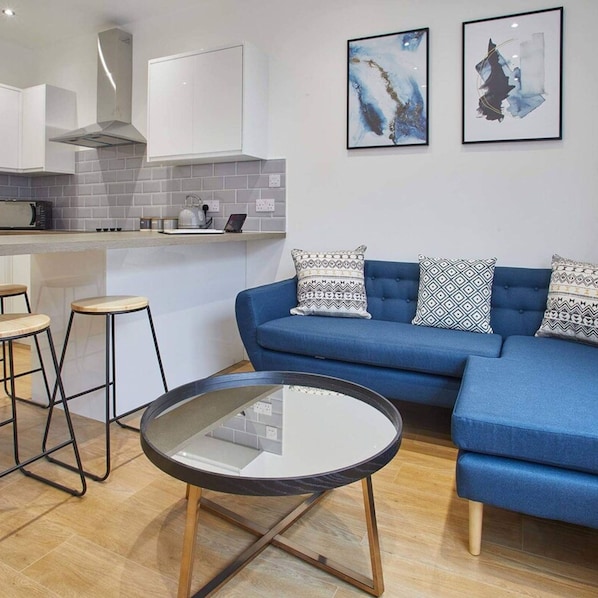 Un salon moderne avec un canapé bleu, deux coussins à motifs, une table basse en verre et un coin cuisine doté de meubles blancs et d’un pare-feu gris.