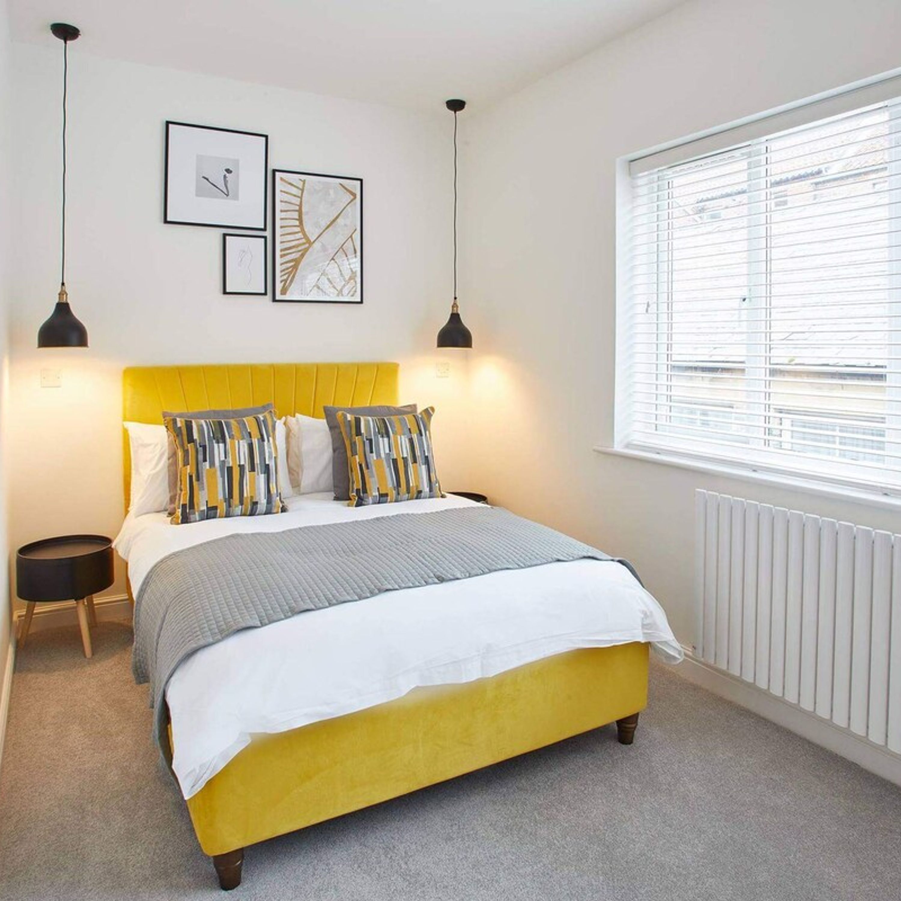 A bedroom with a yellow headboard, a bed with striped bedding, two bedside tables, and a window with blinds.