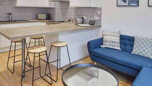 Eine moderne Küche mit weißen Schränken, einer grauen gefliesten Spritzschutzwand und einer zentralen Kochinsel mit Sitzgelegenheiten. Ein blaues Sofa mit gemusterten Kissen und ein Glastisch ergänzen den Raum.