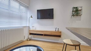 Ein modernes Wohnzimmer mit einem runden Glastisch, zwei hölzernen Hocker, einem Wand-Fernseher, einem Holz-Konsolentisch und einer Pflanze auf einem Wandregal.