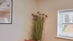 A corner of a room with a beige wall, a framed picture, a tall plant, a window with blinds, and a brown sofa with a cushion. - Host Stay The View Hudsons Yard House (Whitby)