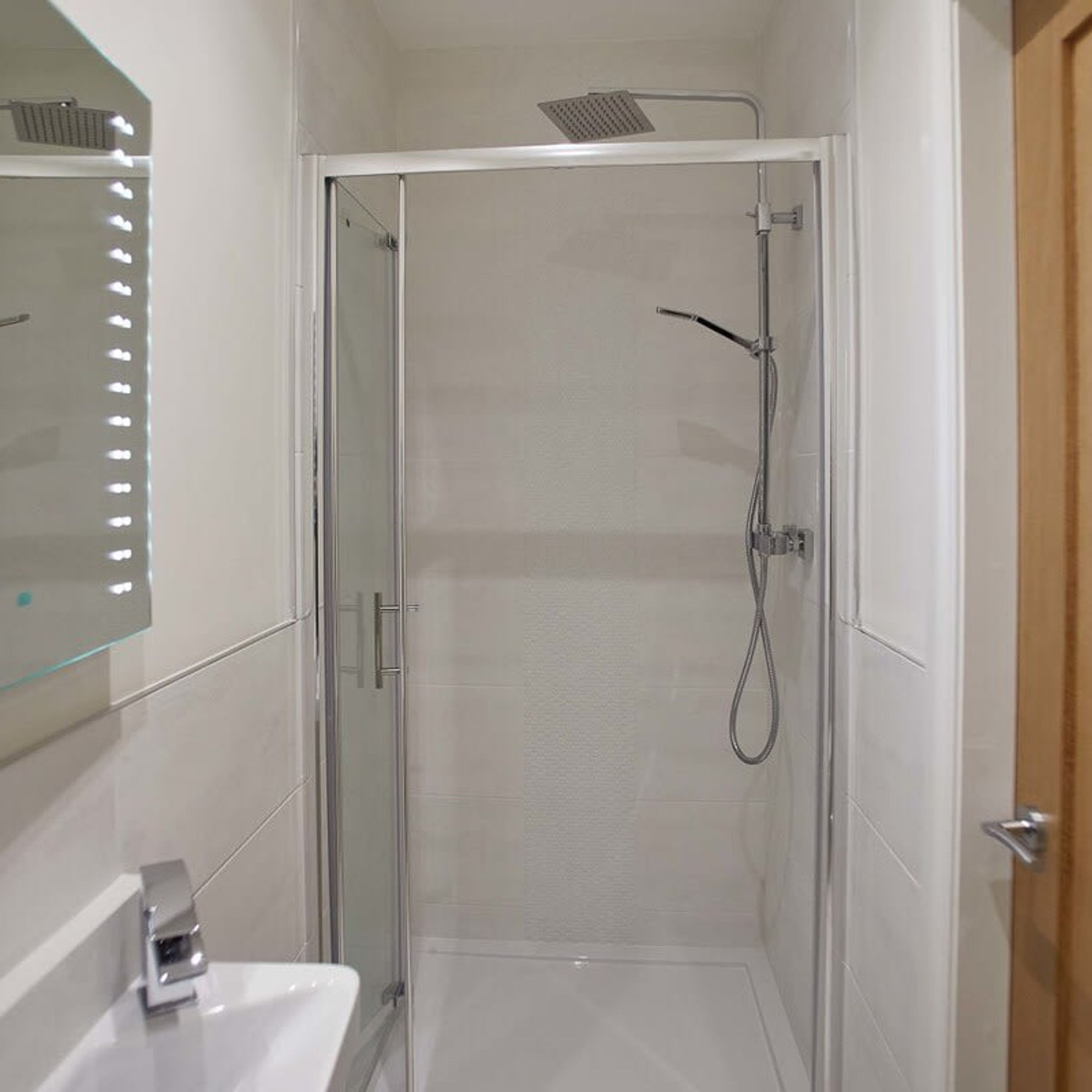 A modern bathroom with a glass shower enclosure, a white sink, and a mirror with lights.