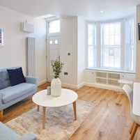 Una sala de estar moderna con un sofá azul claro, una mesa de centro blanca y un sillón blanco. Se observa un radiador, una puerta y una ventana con persianas.