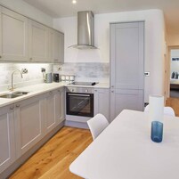 Una cocina moderna con gabinetes grises, una campana extractora de acero inoxidable, un horno empotrado y una mesa de comedor blanca con cubierta de vidrio azul.