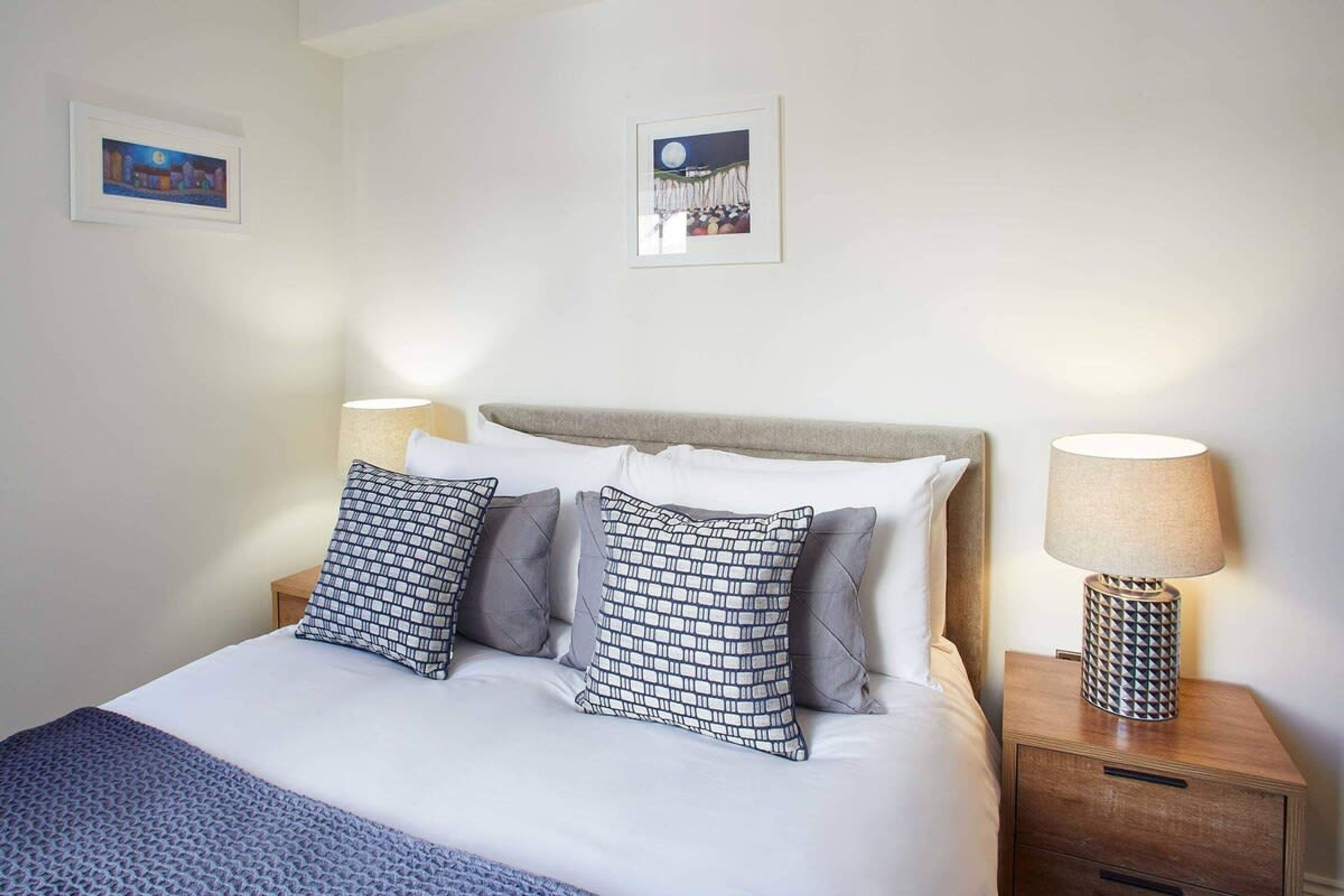 Una cama bien hecha con almohadones estampados, una lámpara de noche y dos cuadros en la pared.