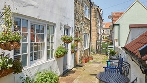 Une cour étroite avec un sol en pierre, des murs blancs et des paniers de fleurs suspendus. On y trouve une table ronde en métal, des chaises et un petit pot bleu contenant des plantes.