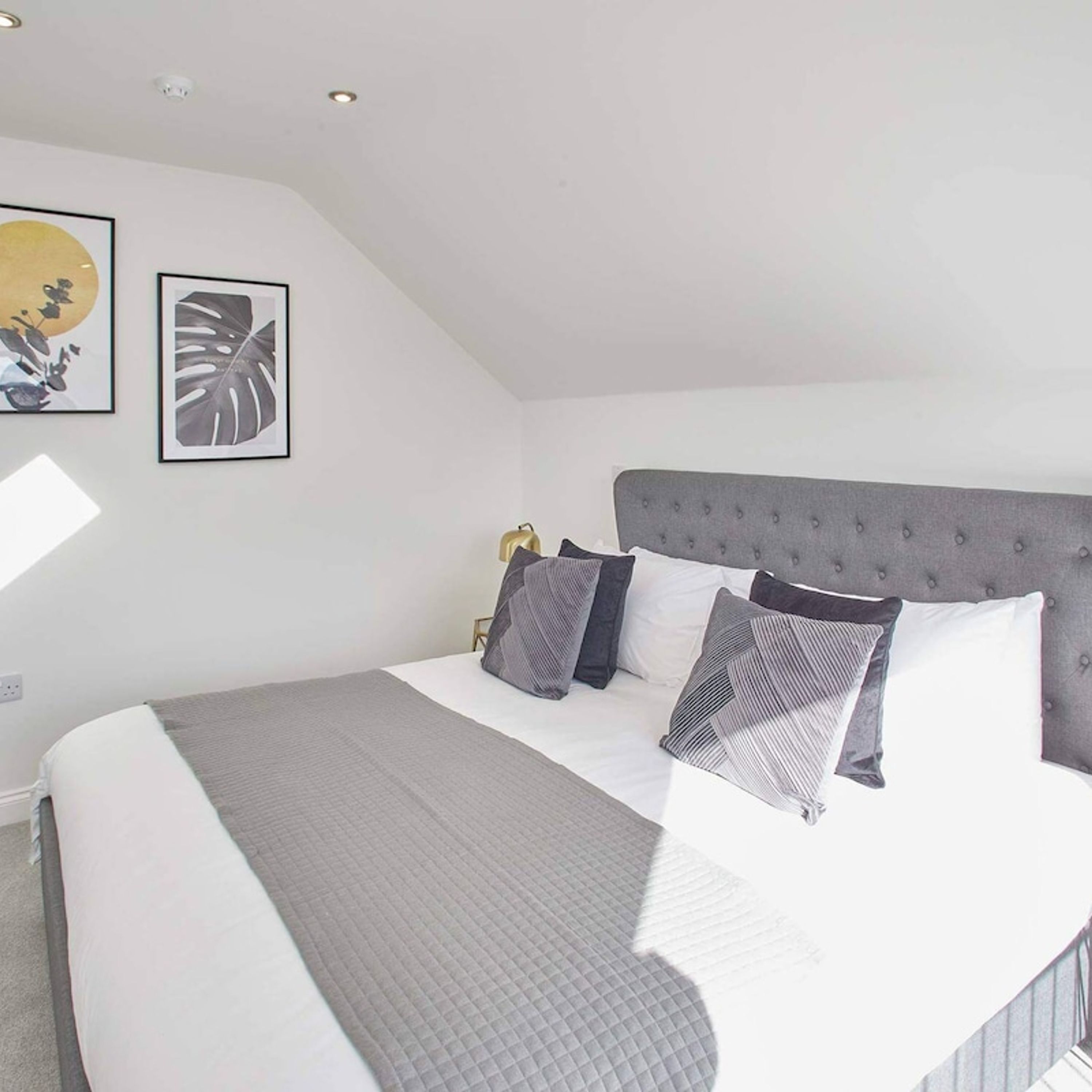 Ein Schlafzimmer mit einem großen Bett, grau-weißer Bettwäsche, einem gepolsterten grauen Kopfteil und zwei gerahmten Kunstwerken an der Wand.