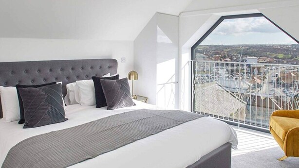A modern bedroom with a large bed, a view of the city through a balcony, and a yellow armchair.