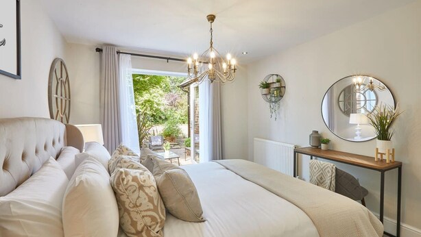 A bedroom with a large bed, a chandelier, a round mirror, and a view of the outdoors. - Host Stay The Hampton at Prospect Hill Apartments (Whitby)
