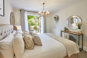 A bedroom with a large bed, a chandelier, a round mirror, and a view of the outdoors. - Host Stay The Hampton at Prospect Hill Apartments (Whitby)