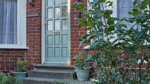 A traditional brick house with a green door, two windows with white frames, and a small porch with potted plants.