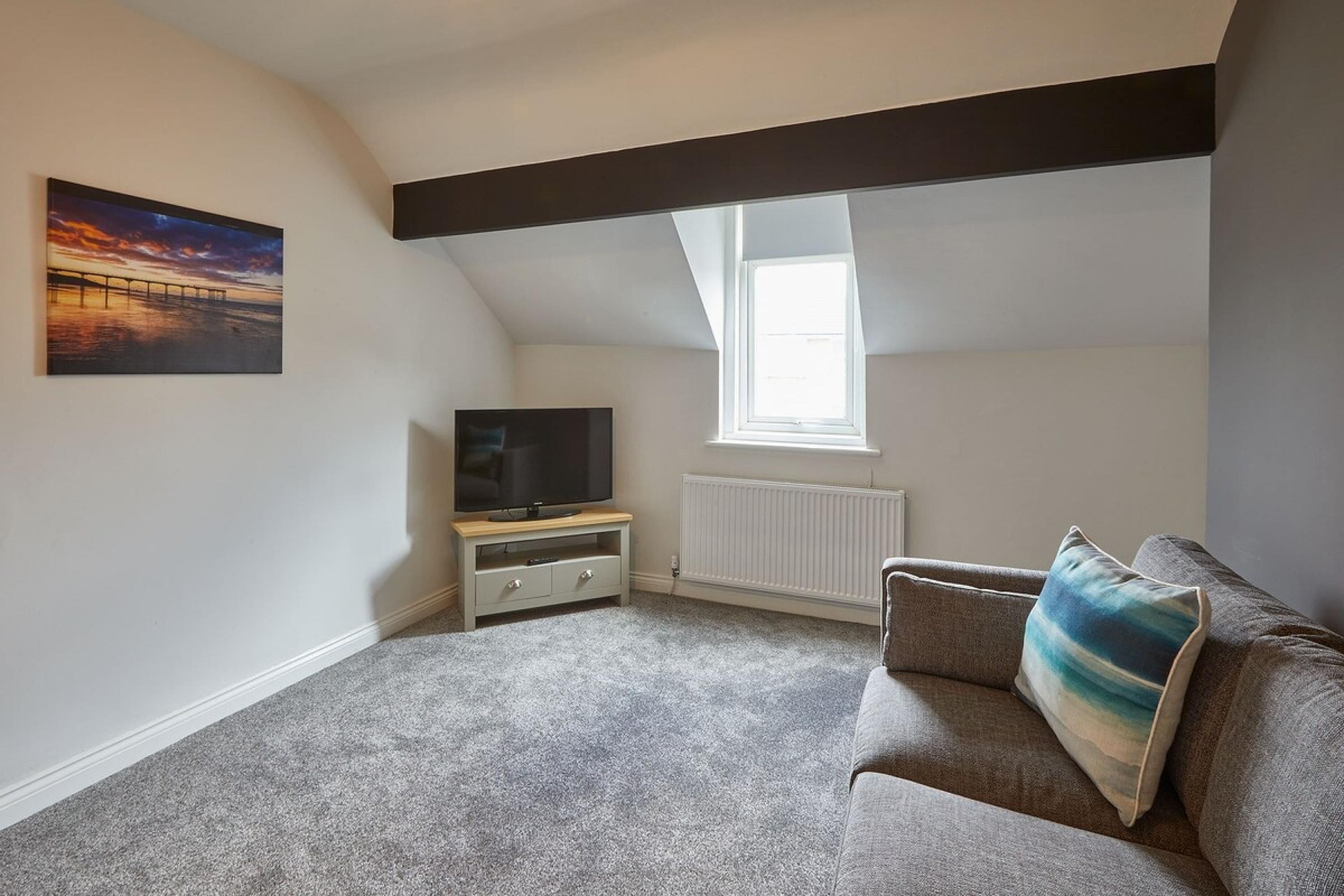 A small, well-lit attic room with a sloped ceiling, a television on a wooden stand, a grey sofa, and a painting of a sunset on the wall.