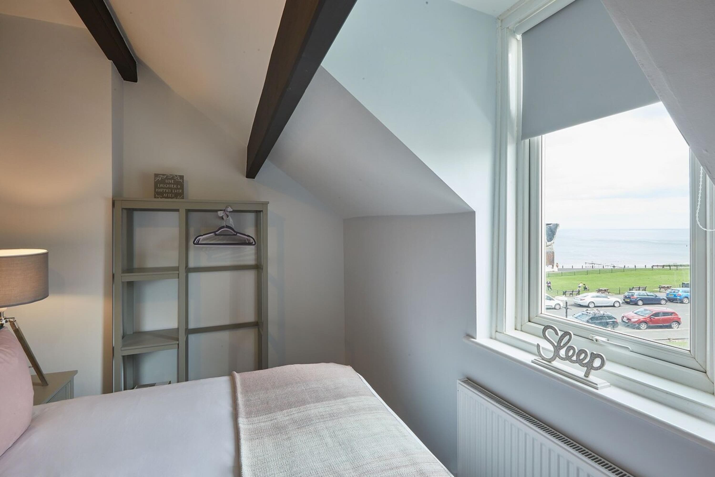 A bedroom with a bed, a hanging rack, and a window with a view of the sea and parked cars.