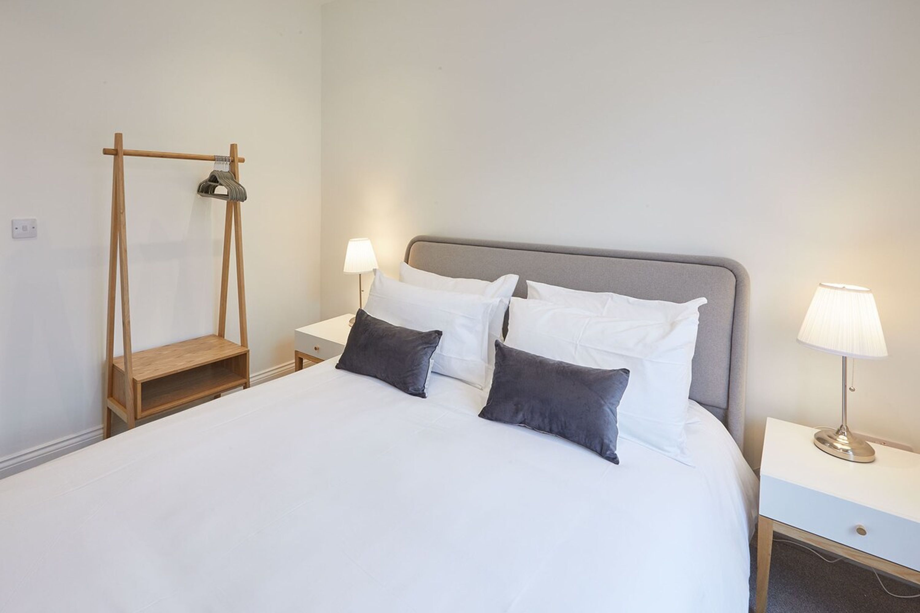 A neatly made bed with white bedding and two gray pillows, a wooden shelf unit, and two bedside lamps on a white nightstand.