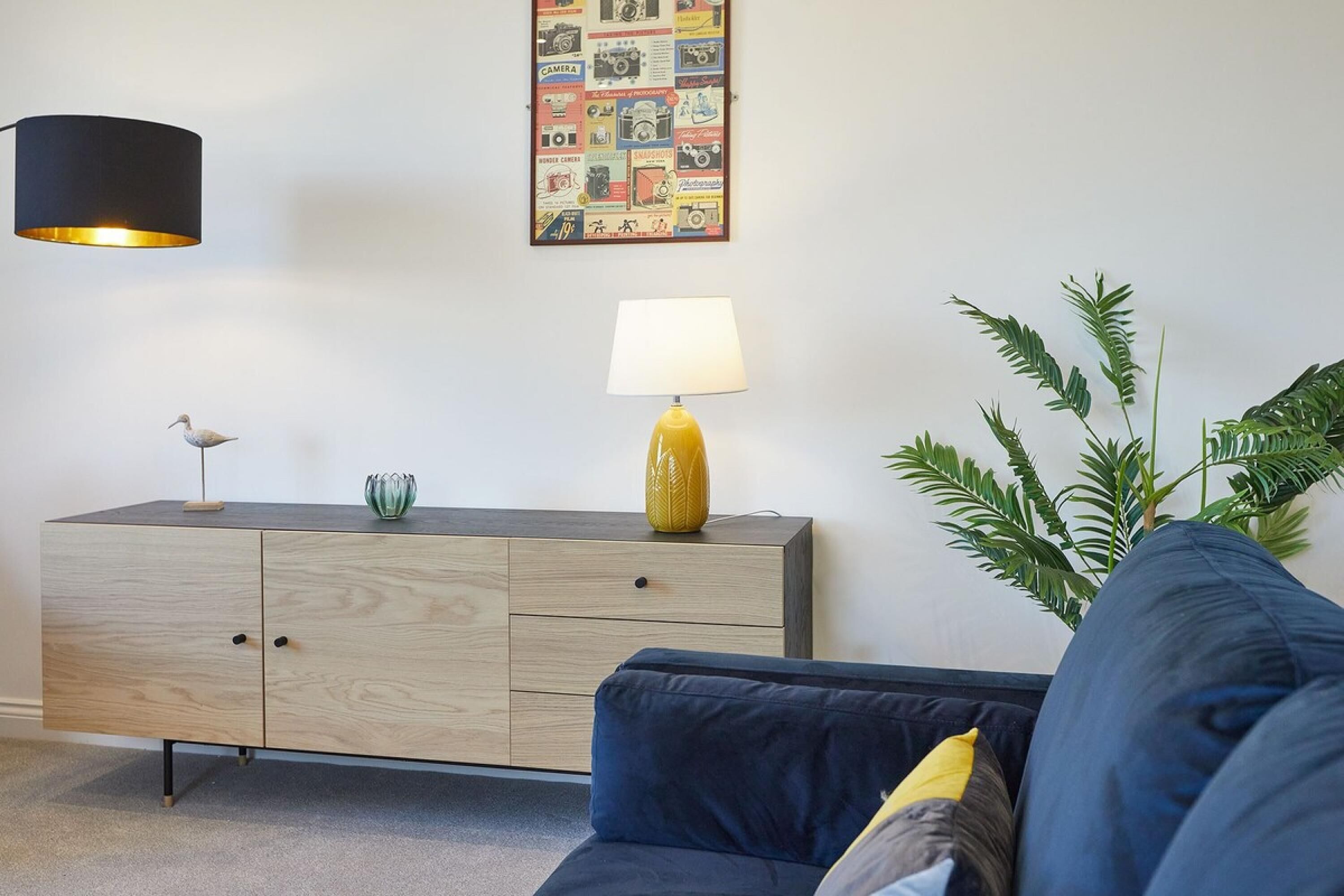 Un salón moderno con un sofá azul oscuro, un mueble de madera con una lámpara amarilla, un cartel de cámara en la pared y una planta verde.