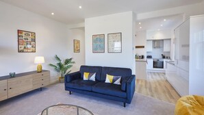 Ein modernes Wohnzimmer mit einem blauen Sofa, einem Glastisch und einem gelben Sitzsack. Im Hintergrund ist eine Küche mit weißen Schränken und einem Herd zu sehen.