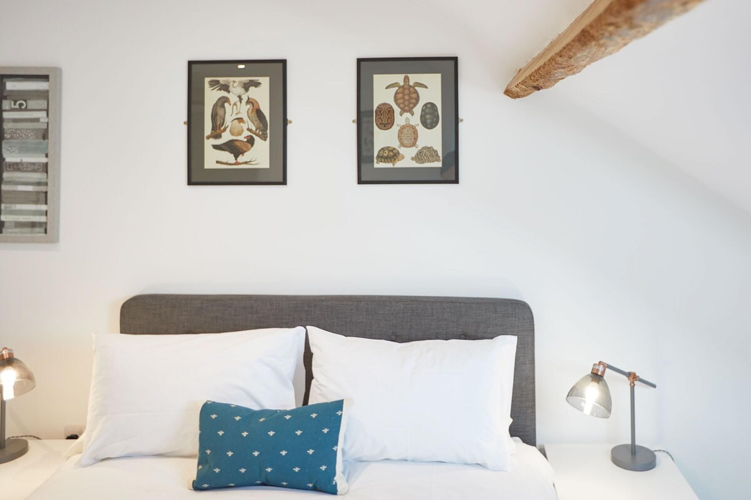 A bedroom with a bed, two framed pictures on the wall, a nightstand with a lamp, and a wooden beam above the bed.