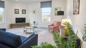 Ein modernes Wohnzimmer mit einer Couch, einem Fernseher, einem Couchtisch und einem Sessel. Es gibt Fenster mit Jalousien, eine Heizung und eine Pflanze.