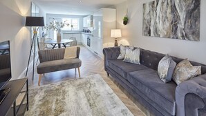 A modern living room with a grey sofa, a coffee table, and a floor lamp. There is a dining area with a table and chairs in the background. - Host Stay The Madison at Prospect Hill Apartments (Whitby)