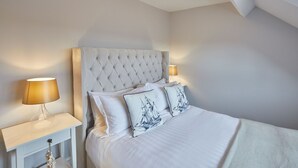 A bedroom with a tufted headboard, white bedding, two matching pillows, a bedside table with a lamp, and a nightstand with a decorative item. - Host Stay Henderson House (Whitby)