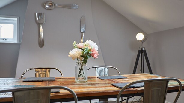 A dining area with a wooden table, metal chairs, a vase with flowers, and wall-mounted light fixtures. - Host Stay Henderson House (Whitby)