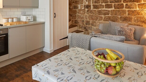 A cozy kitchen with white cabinets, a stone wall, and a dining area with a table set with a fruit bowl. - Host Stay Dolphin Cottage 1736 (Whitby)