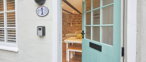 A teal door labeled "Dolphin Cottage," a stone wall, a white doorbell, and a window with blinds.