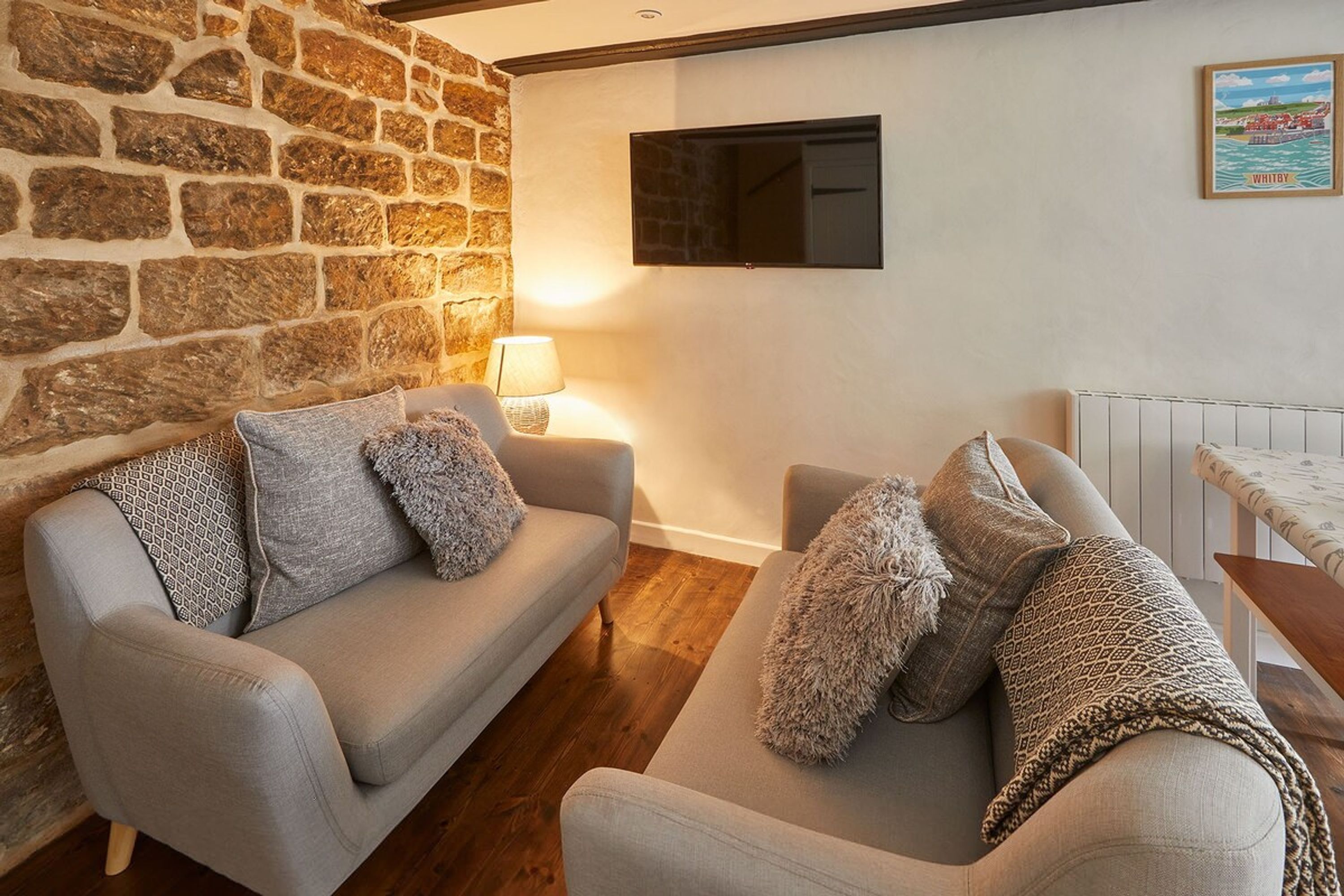 A cozy living room with a stone wall, two sofas with patterned cushions, a wooden floor, a mounted TV, and a framed picture on the wall.