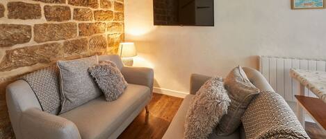 A cozy living room with a stone wall, two sofas with patterned cushions, a wooden floor, a mounted TV, and a framed picture on the wall.