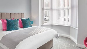 Ein Schlafzimmer mit einem Bett, einem Fenster mit Jalousien, einem blauen Teppich und einem rosa Sessel.